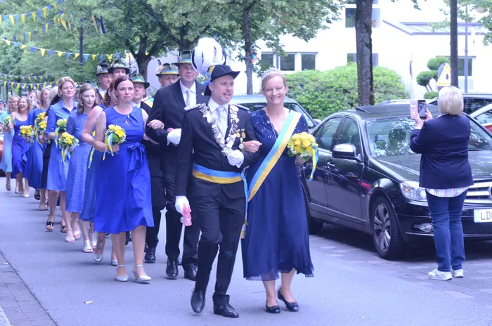 Ein Paar in festlicher Tracht geht vor einer Gruppe von Menschen her, die an einem Umzug teilnehmen. Im Hintergrund sind Gebäude und ein schwarzer Wagen zu sehen.