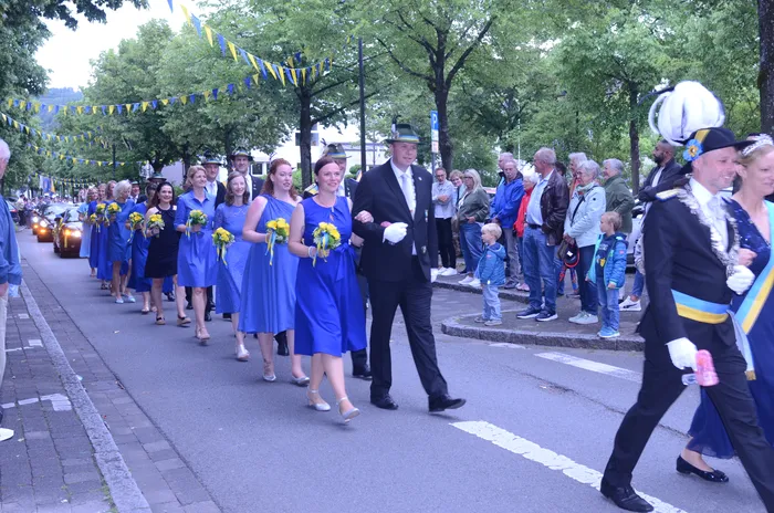 Eine Gruppe von Menschen schreitet in einem Festzug die Straße entlang. Die Frauen tragen blaue Kleider und halten gelbe Blumensträuße, während der Mann in der Mitte einen schwarzen Anzug trägt. Am Straßenrand stehen Zuschauer und beobachten den Umzug.