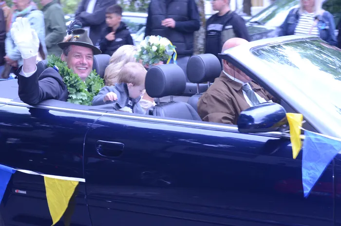 Ein Mann in einem dunklen Anzug und Hut winkt aus einem offenen Auto, während er an einer Menschenmenge vorbeifährt. Im Hintergrund sind weitere Personen und blau-gelbe Flaggen zu sehen.