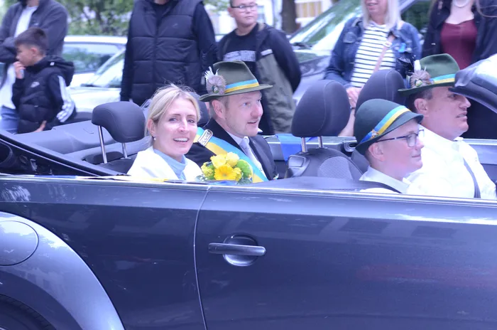 Drei Männer und eine Frau sitzen lächelnd in einem offenen schwarzen Wagen. Die Frau hält einen Blumenstrauß, die Männer tragen grüne Hüte mit Federn und Uniformen. Im Hintergrund sind weitere Menschen zu sehen.