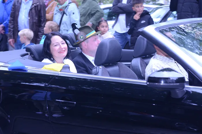 Drei Personen sitzen in einem offenen schwarzen Cabriolet und lächeln in die Kamera. Die Frau links trägt ein gelbes Oberteil, der Mann in der Mitte einen grünen Hut mit Federn. Im Hintergrund sind Zuschauer, darunter Kinder, zu sehen.