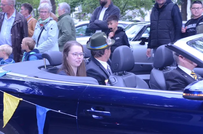 Ein Cabriolet fährt an einer Menschenmenge vorbei. Im Auto sitzen Personen in festlicher Kleidung, darunter ein Mann mit einem grünen Hut und eine junge Frau mit Brille. Im Hintergrund stehen und sitzen weitere Zuschauer.