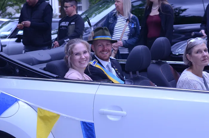 A man with a beard wearing a hat and uniform smiles from the passenger seat of a white convertible, alongside a young woman in a light pink dress. A woman is visible in the back seat. The car is decorated with blue and yellow ribbons.