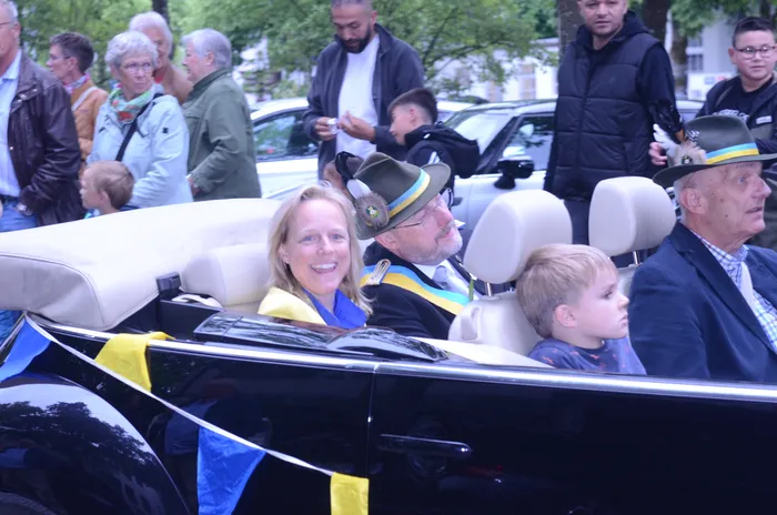 Eine Frau lächelt in einem offenen schwarzen Wagen neben einem Mann in einem grünen Hut mit Abzeichen. Im Fond sitzt ein Junge, während im Hintergrund Zuschauer stehen.
