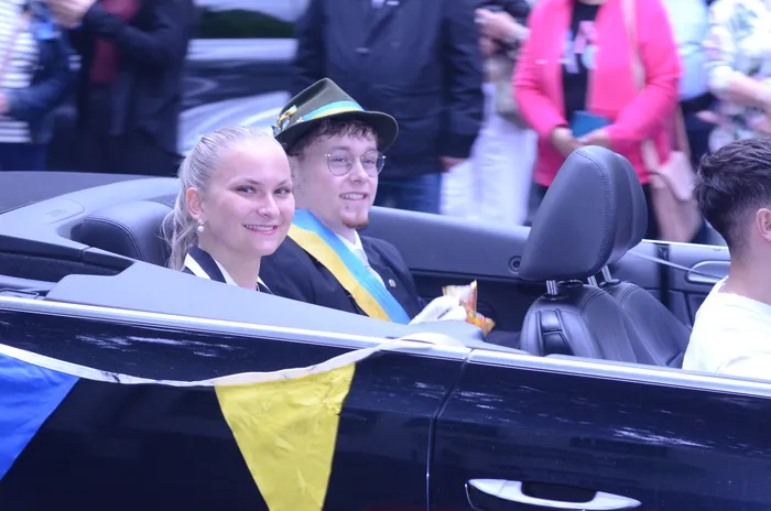 Eine Frau und ein Mann sitzen lächelnd in einem offenen Cabriolet. Der Mann trägt einen grünen Hut und ein blau-gelbes Band, während die Frau einen dunklen Anzug trägt. Im Hintergrund sind Menschen und ein blau-gelbes Banner zu sehen.