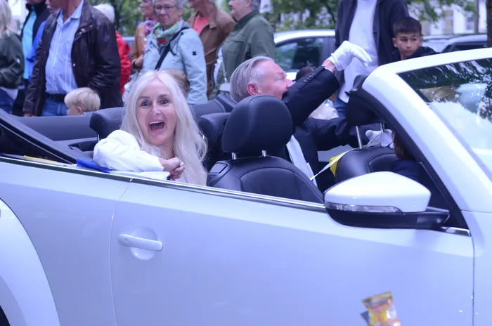 Eine Frau mit langen blonden Haaren und ein Mann im offenen weißen Cabriolet winken in die Menge. Im Hintergrund stehen Menschen und beobachten den Festzug.