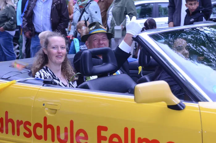 A woman with curly hair and a man in a hat and blue shirt wave from a yellow convertible during a parade. The car is labeled 'Fahrschule Follmer'.