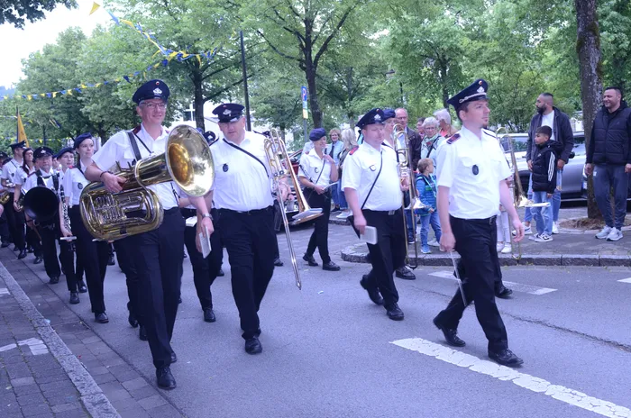 Eine Blaskapelle marschiert auf einer Straße, die Musiker tragen weiße Uniformen und dunkle Hosen. Im Hintergrund sind Zuschauer und Bäume zu sehen.