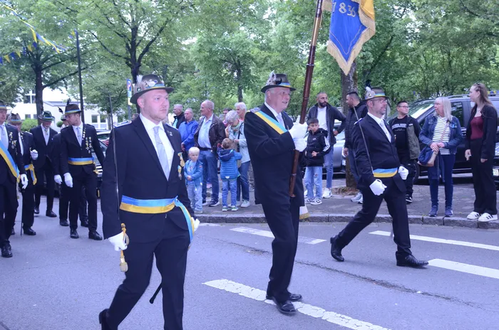 Eine Gruppe von Männern in dunklen Uniformen und mit Zylindern marschiert auf einer Straße, einige tragen Fahnen. Im Hintergrund stehen Zuschauer, darunter auch Kinder und Frauen.