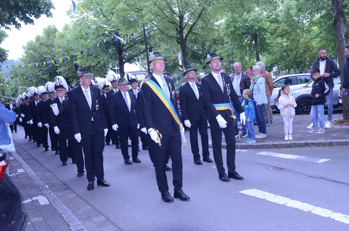 Eine Gruppe von Männern in dunklen Anzügen, weißen Handschuhen und Hüten marschiert auf einer Straße. Einige tragen Schärpen und halten Degen. Zuschauer stehen am Straßenrand.