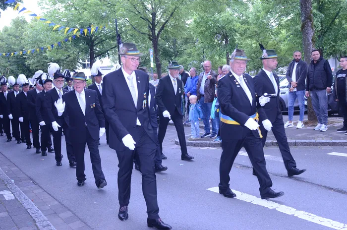 Eine Gruppe von Männern in dunklen Anzügen, weißen Handschuhen und Hüten marschiert auf einer Straße. Einige tragen Abzeichen und Kokarden. Im Hintergrund stehen Zuschauer und geparkte Autos.