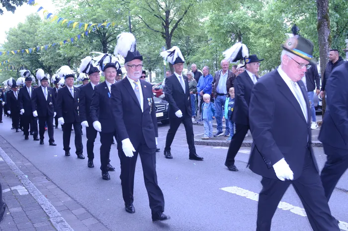 Eine lange Reihe von Männern in dunklen Anzügen und mit weißen Handschuhen marschiert auf einer Straße, einige tragen Federhüte. Zuschauer stehen am Straßenrand und beobachten den Festzug.