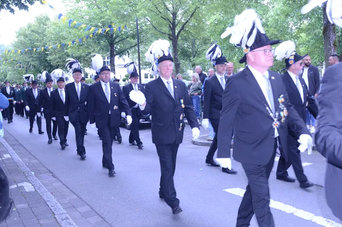 Eine Gruppe von Männern in dunklen Anzügen und mit weißen Federn auf ihren Hüten marschiert auf einer Straße. Sie tragen weiße Handschuhe und einige tragen Orden. Im Hintergrund sind Zuschauer und Girlanden zu sehen.
