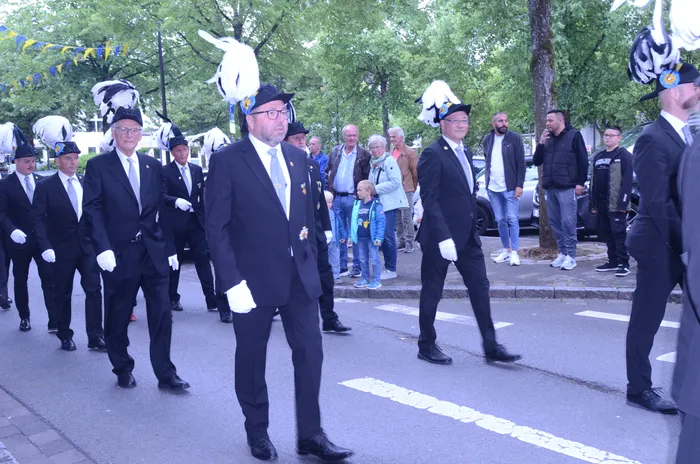 Eine Gruppe von Männern in dunklen Anzügen und mit weißen Federbüschen auf ihren Hüten marschiert auf einer Straße. Einige Zuschauer stehen am Straßenrand und beobachten den Festzug.