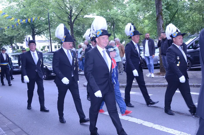 Männer in dunklen Anzügen und mit hohen, weißen Federbüschen auf den Hüten marschieren in einer Reihe auf einer Straße. Einige tragen weiße Handschuhe und Abzeichen. Im Hintergrund sind Zuschauer und Gebäude zu sehen.