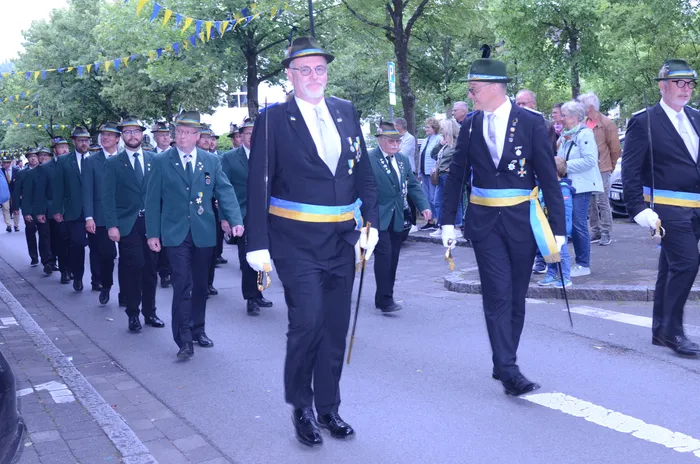 Eine Gruppe von Männern in dunklen Anzügen und Zylindern marschiert auf einer Straße, einige tragen Orden und halten Spazierstöcke. Im Hintergrund sind Zuschauer und Girlanden zu sehen.