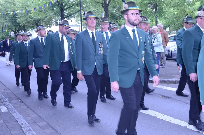 Eine Gruppe von Männern in grünen Jacken, schwarzen Hosen und Hüten marschiert auf einer Straße. Viele tragen Abzeichen und Medaillen auf ihren Jacken. Im Hintergrund sind weitere Personen und ein Auto zu sehen.