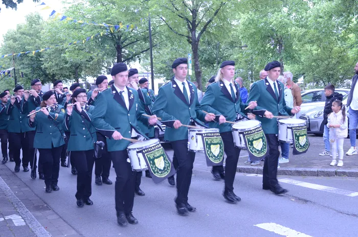 Eine Gruppe von jungen Männern und Frauen in grünen Uniformen und schwarzen Hüten marschiert auf einer Straße. Einige spielen Flöten, andere tragen große Trommeln mit dem Aufdruck 