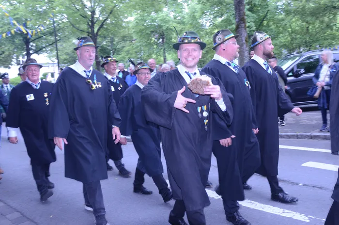 Eine Gruppe von Männern in dunklen Anzügen und Zylinderhüten marschiert auf einer Straße. Einige tragen Orden und ein Mann hält eine Brezel hoch.