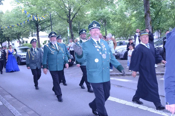 Eine Gruppe von Männern in traditionellen grünen Uniformen und Hüten marschiert auf einer Straße, während ein Mann im Vordergrund die Hand hebt. Im Hintergrund sind weitere Menschen und Autos zu sehen.