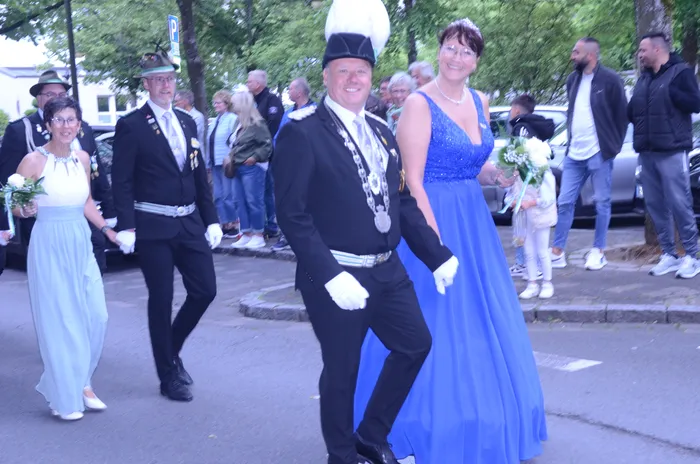 Ein Mann in schwarzem Festzugsgewand mit weißem Handschuh und eine Frau in blauem Abendkleid gehen lächelnd durch eine Menschenmenge. Im Hintergrund sind weitere Festteilnehmer und Zuschauer zu sehen.