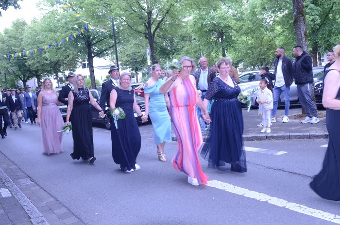 Eine Gruppe von Menschen, darunter Frauen in Abendkleidern, geht auf einer Straße in einer städtischen Umgebung. Einige halten Blumensträuße. Im Hintergrund sind weitere Menschen und Bäume zu sehen.