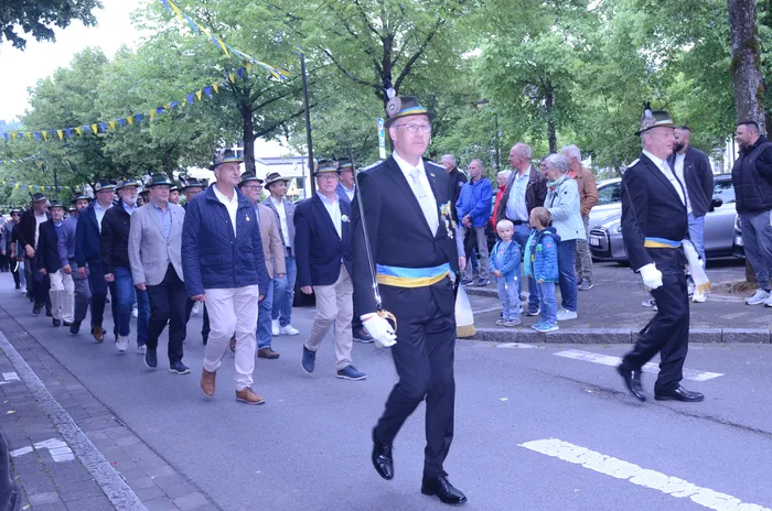 Eine Gruppe von Männern in verschiedenen Anzügen und Uniformen marschiert auf einer Straße, einige tragen Hüte und weiße Handschuhe. Im Hintergrund sind Zuschauer und Girlanden zu sehen.