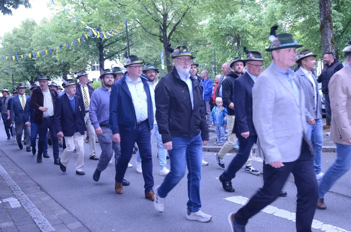 Eine Gruppe von Männern marschiert in einer Reihe die Straße entlang. Die meisten tragen traditionelle Hüte mit Federn und formelle Kleidung. Im Hintergrund sind Bäume und festliche Girlanden zu sehen.