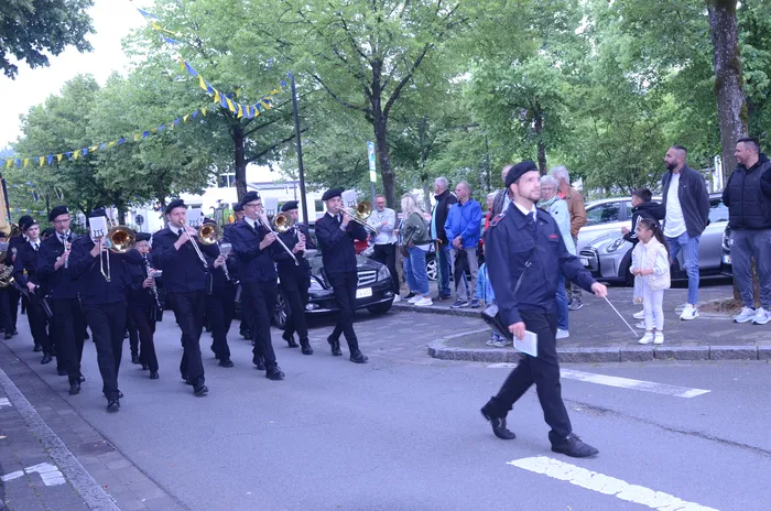 Eine Marschkapelle in dunklen Uniformen spielt auf, während sie an einer Straße entlangzieht. Zuschauer stehen am Straßenrand und beobachten den Festzug.
