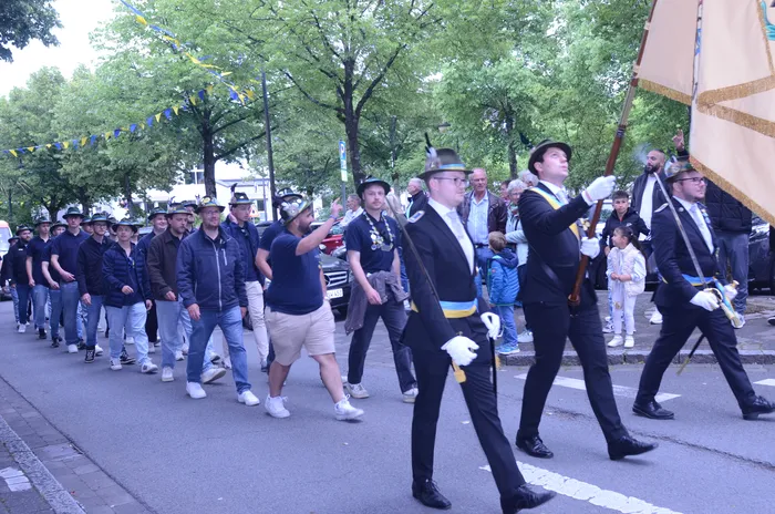 Eine Gruppe von Männern in dunklen Anzügen und Zylindern marschiert auf einer Straße, einige tragen Fahnen. Im Hintergrund sind weitere Menschen und Bäume zu sehen.