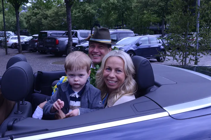 Familie im Cabriolet während des Schützenfestumzugs. Ein Mann mit Hut lächelt in die Kamera, neben ihm ein Kind und eine Frau. Im Hintergrund geparkte Autos.