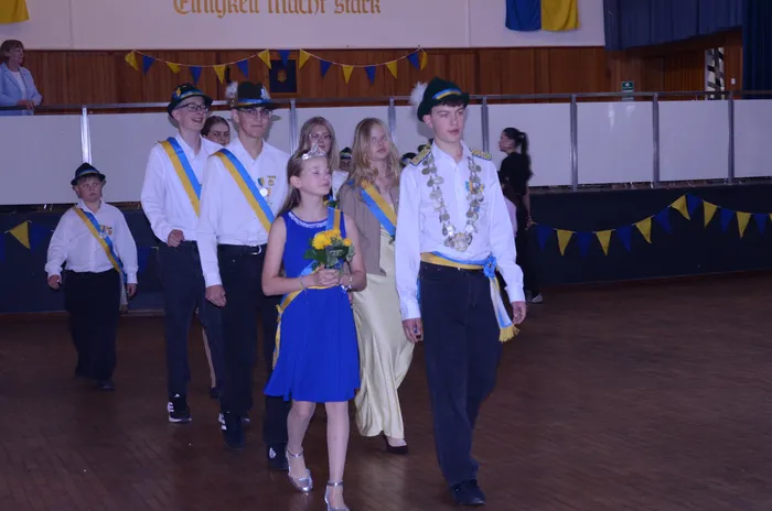 Eine Gruppe von Jugendlichen und Kindern in festlicher Kleidung schreitet durch einen Raum. Einige tragen traditionelle Schützenuniformen mit Hüten und Bändern, andere sind in Kleidern und Anzügen gekleidet. Im Hintergrund sind Wimpelketten und ein Banner zu sehen.