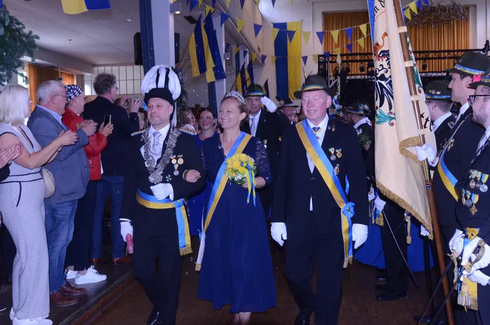 Ein Paar in festlicher Tracht mit Orden und blauem Band schreitet durch eine Menschenmenge. Im Hintergrund sind Girlanden und weitere Personen in Uniformen zu sehen.