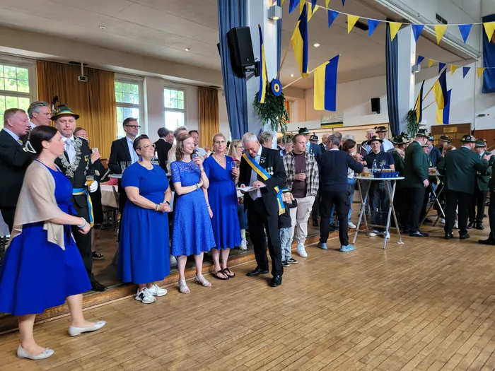Menschen in formeller Kleidung stehen und unterhalten sich in einem hellen Raum mit Holzboden. Einige Frauen tragen blaue Kleider, während Männer Anzüge oder Uniformen tragen. Im Hintergrund sind Girlanden und Dekorationen zu sehen.