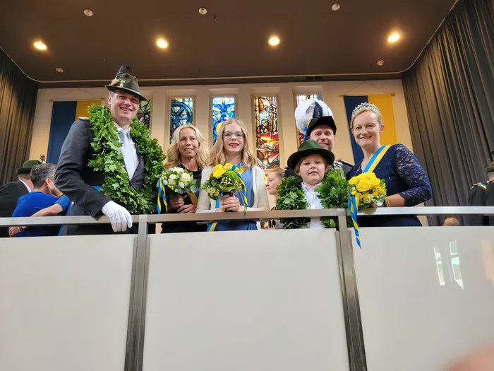 Eine Gruppe von sechs Personen steht auf einer erhöhten Plattform mit Geländer. Die Gruppe besteht aus vier Erwachsenen und zwei Kindern. Die Erwachsenen tragen traditionelle Trachten und Schützenabzeichen, einige halten Blumensträuße. Im Hintergrund sind Buntglasfenster und ein Raum mit Zuschauern zu sehen.