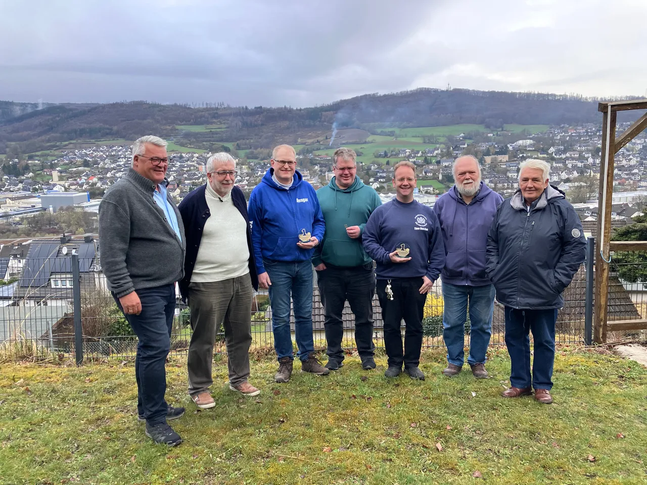 Sieben Männer mittleren Alters stehen lächelnd auf einer Anhöhe vor einer Stadtlandschaft. Einige halten kleine Pokale oder Figuren in den Händen, vermutlich Auszeichnungen für sportliche Leistungen.