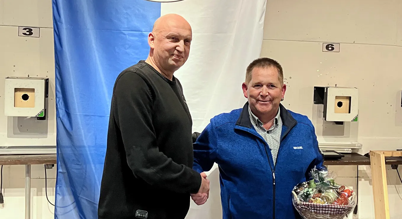 Zwei Männer im mittleren Alter stehen sich lächelnd gegenüber und geben sich die Hand. Im Hintergrund sind Schießscheiben und eine blau-weiße Fahne zu sehen.