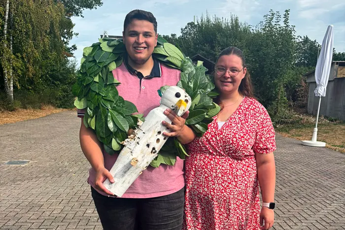 Marcel Schmellenkamp und Lisa Kahl stehen im Freien; Marcel hält einen großen grünen Kranz und eine weiße Holzfigur.