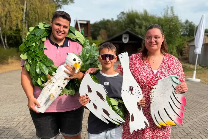 Drei junge Menschen halten große, weiße Vogelfiguren; der junge Mann links trägt einen grünen Laubkranz.