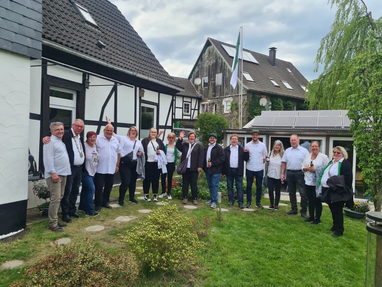 Eine Gruppe von etwa zwanzig Personen steht lächelnd vor einem Fachwerkhause mit Solarzellen auf dem Dach. Viele halten Biergläser oder -flaschen in der Hand.
