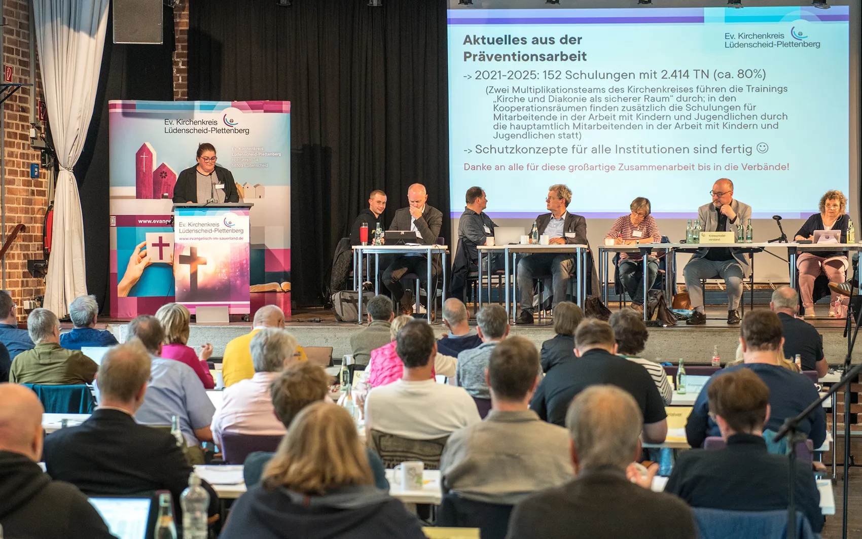 Ein Podium aus mehreren Personen diskutiert vor einem Publikum in einem hellen Raum. Im Hintergrund ein Banner mit dem Logo des Ev. Kirchenkreises Lüdenscheid-Plettenberg und Informationen zu Schulungen zum Thema Prävention.