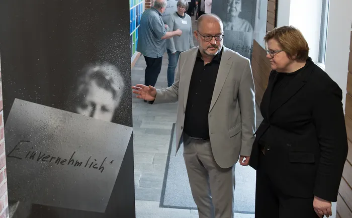 Zwei Personen betrachten ein großes, dunkles Bild mit dem Wort „Einvernehmlich“ darauf. Der Mann trägt einen hellgrauen Anzug und deutet auf das Bild, während die Frau mit Brille ihm aufmerksam zuhört. Im Hintergrund sind weitere Personen und Bilder zu sehen.