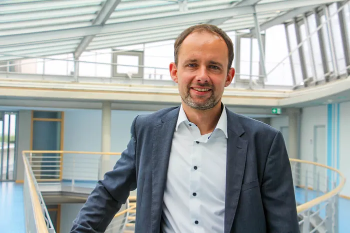 Sebastian Unkhoff lächelt in einem hellen Gebäude mit Glasdach und Metallgeländer.