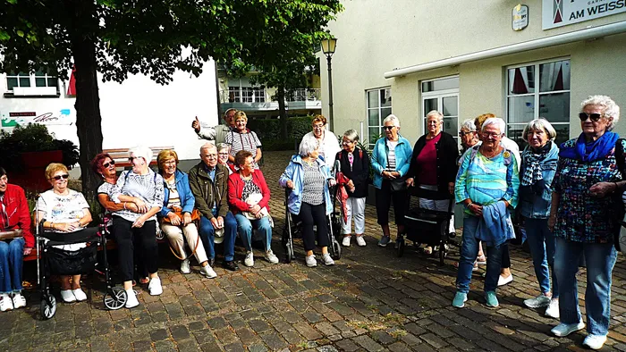 Eine große Gruppe von Senioren posiert für ein Gruppenfoto auf einem gepflasterten Platz vor einem hellen Gebäude.