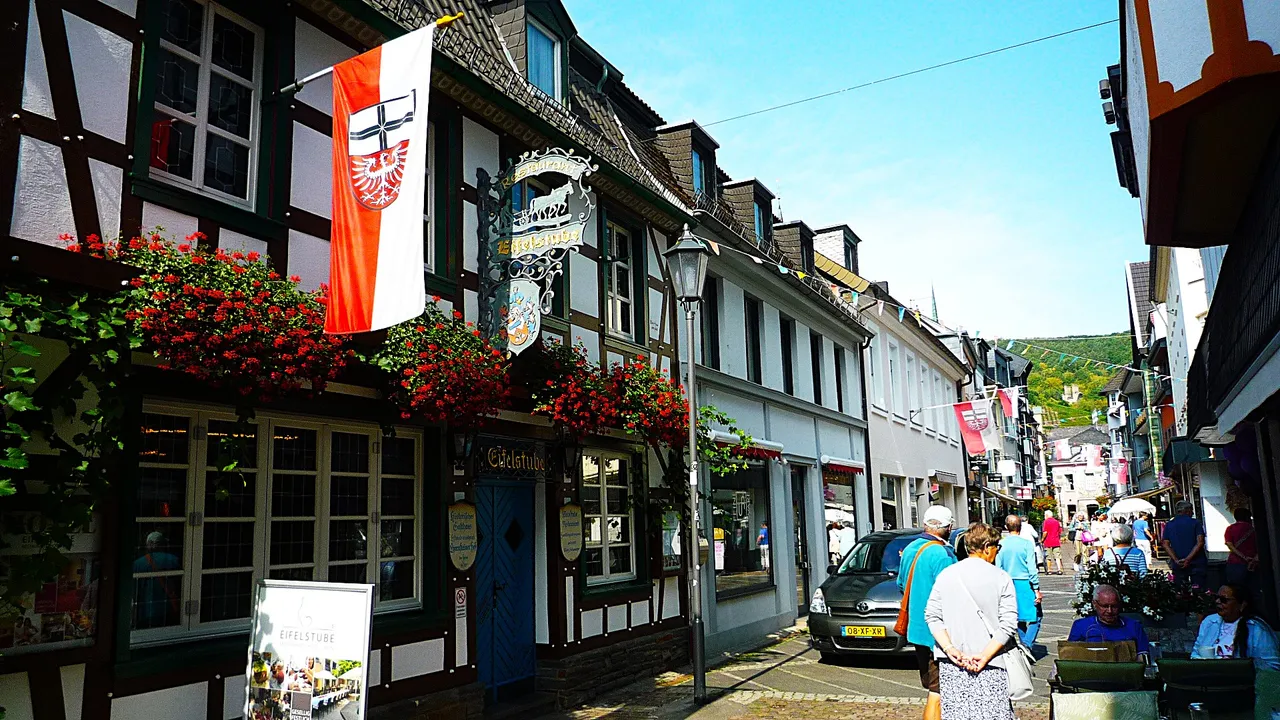 Eine sonnige Straße in der historischen Altstadt von Ahrweiler mit Fachwerkhäusern, blühenden Blumen und einer rot-weißen Flagge.