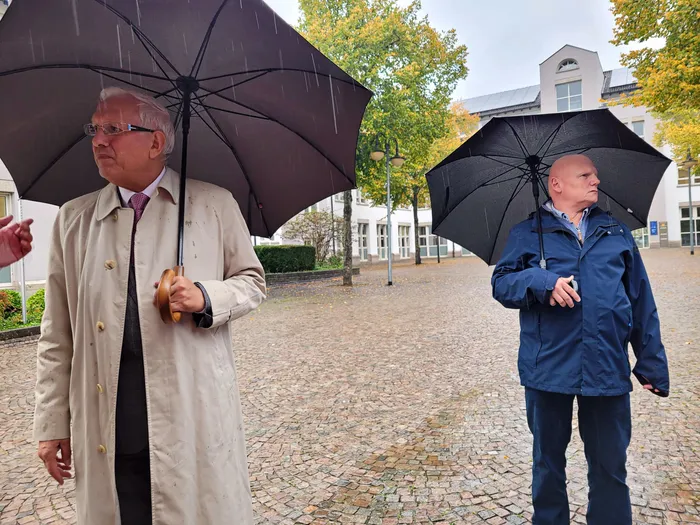 Bürgermeister Ralf Beßler und Stadtplaner Till Hoffmann gehen bei Regen mit schwarzen Regenschirmen über einen gepflasterten Platz.