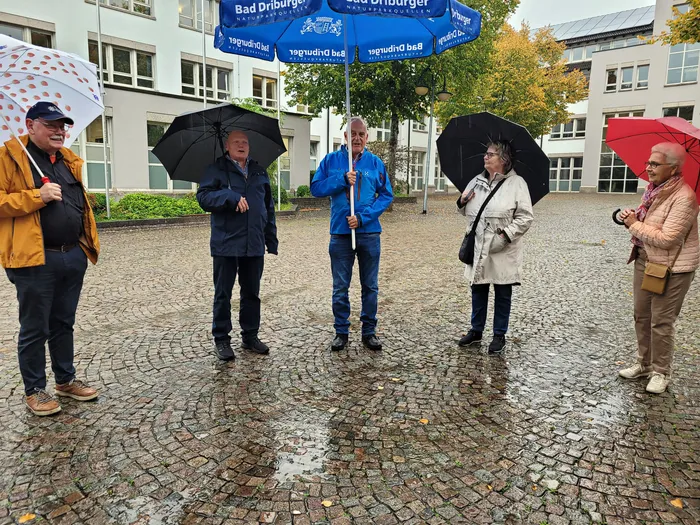 Fünf Personen mit Regenschirmen stehen bei Regen auf einem gepflasterten Platz während einer Innenstadtbegehung.