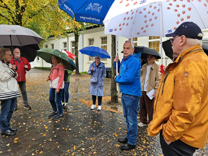 Eine Gruppe von Menschen steht unter Regenschirmen auf einem nassen, mit Blättern bedeckten Kopfsteinpflaster bei einer Innenstadtbegehung.