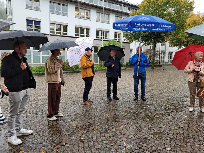 Mehrere Personen mit Regenschirmen stehen auf einer nassen Kopfsteinpflasterstraße vor einem Gebäude während einer Innenstadtbegehung.