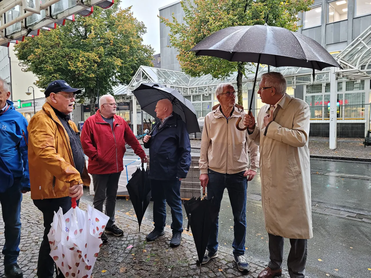 Eine Gruppe von Männern steht bei Regen mit Regenschirmen auf einer nassen, gepflasterten Straße während einer Innenstadtbegehung.